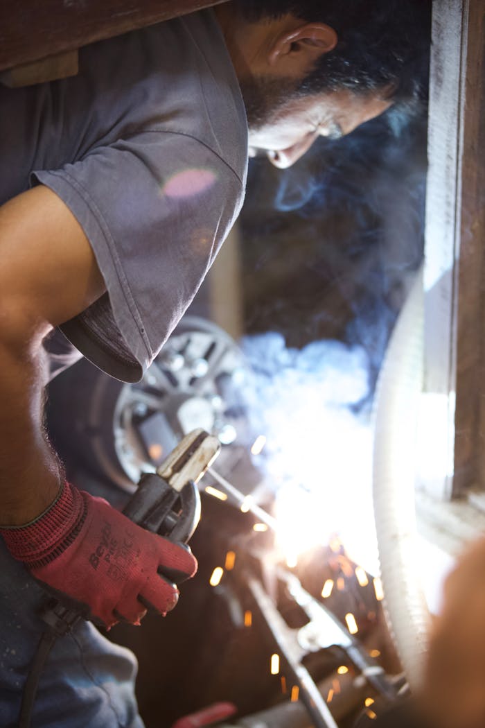 gallery-5 A welder wearing gloves intensely concentrating on welding metal in a workshop setting.