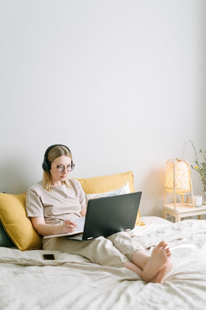 gallery-2 A young woman with headphones works on a laptop in a bright and cozy bedroom setting.