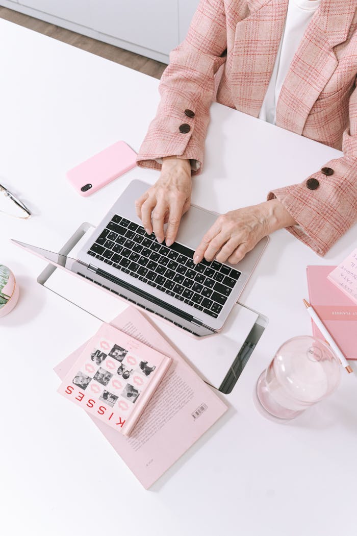 hero-img-02 An adult typing on a laptop in a stylish pink-themed workspace with books and gadgets.