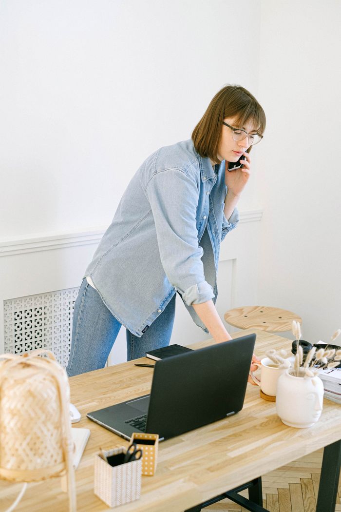 ours-journey Woman multitasking with phone and laptop in bright home office setting.