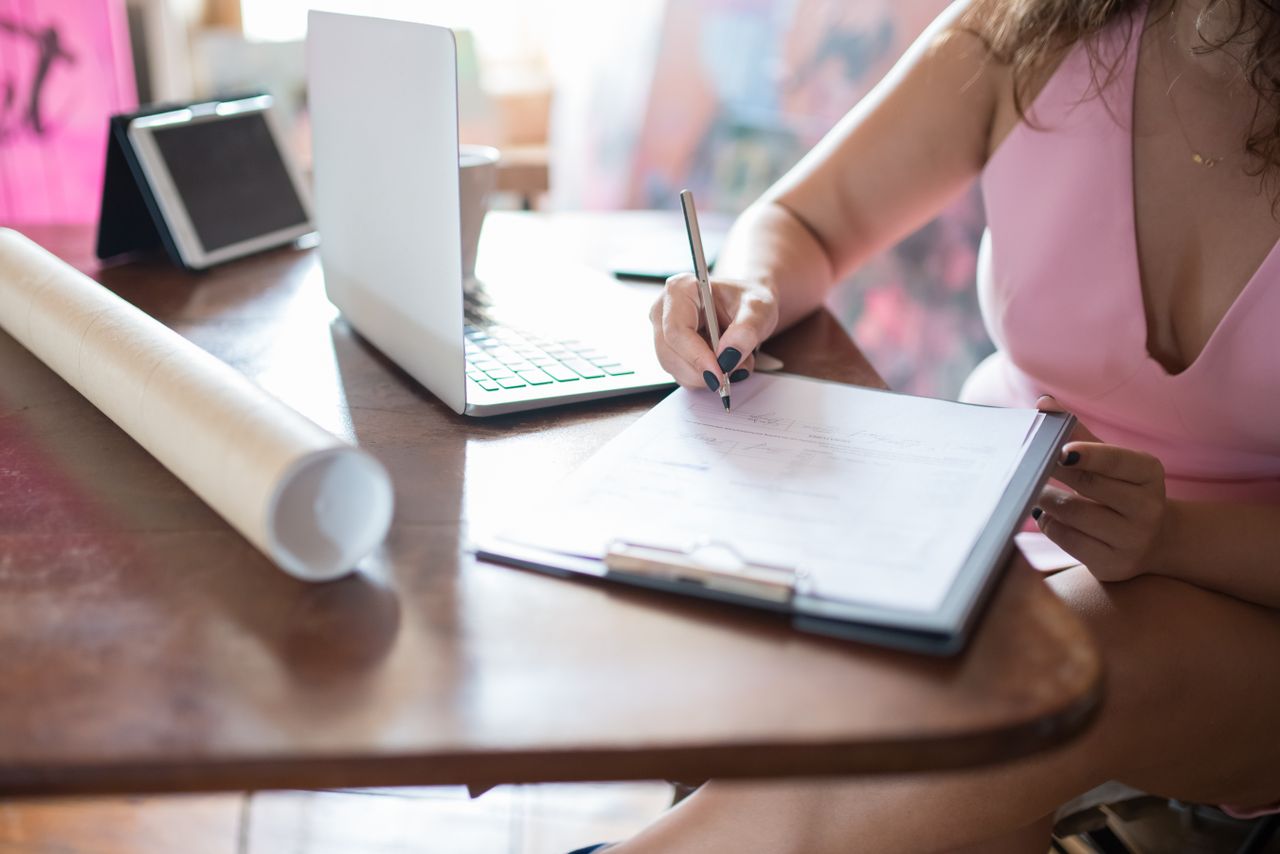 about-img Businesswoman writing on a clipboard at a table with a laptop indoors.