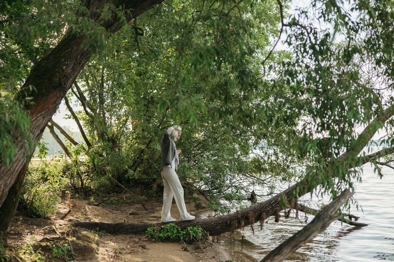 gallery-3 An elderly woman gracefully balances on a fallen tree trunk by a serene lakeside surrounded by lush greenery.